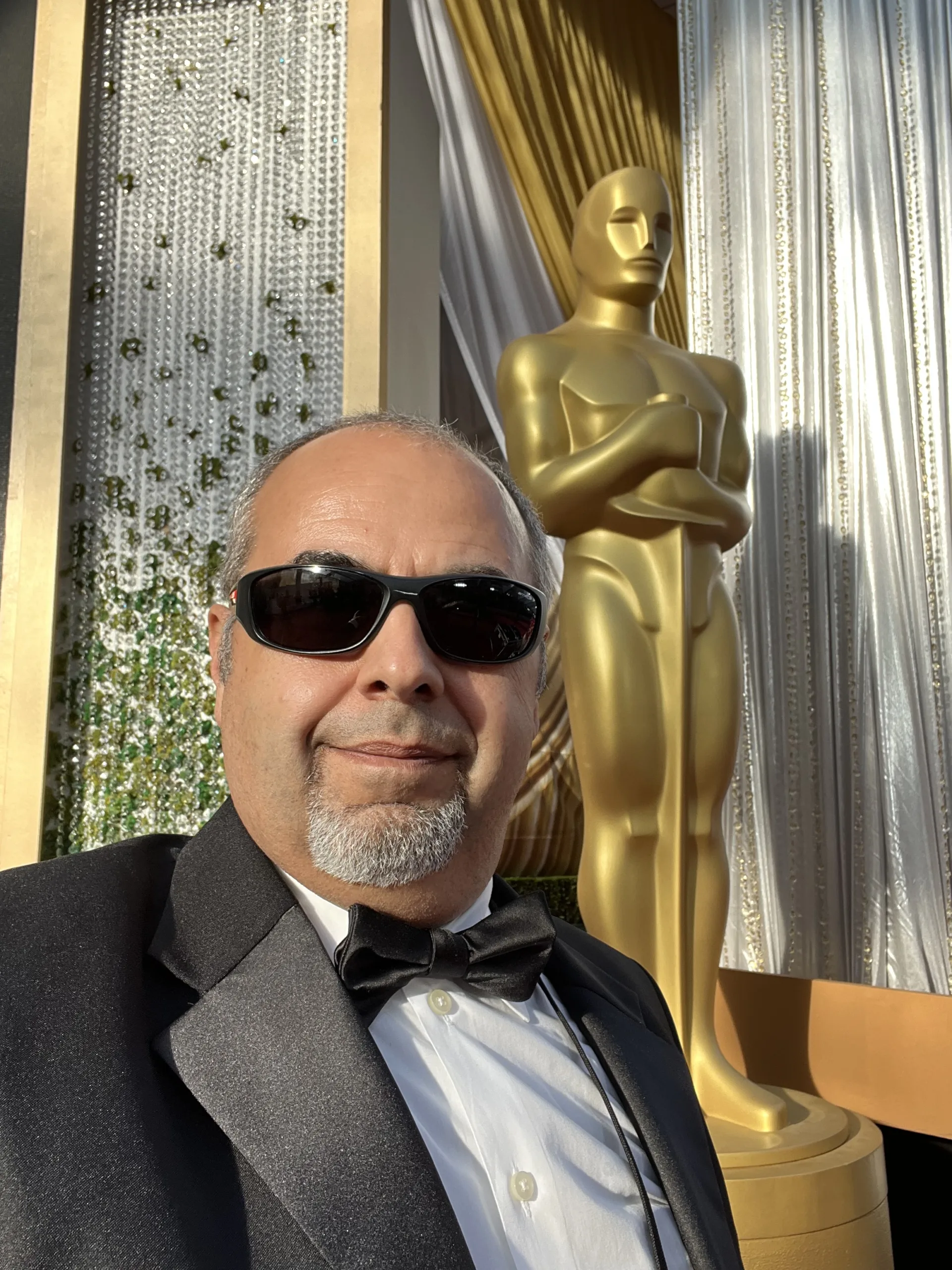 Photo Mar 27 2022, 5 34 35 PM A man wearing sunglasses and a tuxedo in front of an oscar statue.